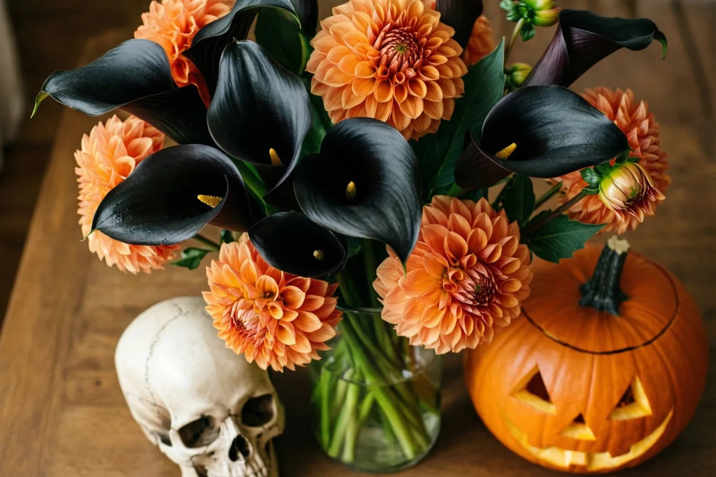 Een glazen vaas met zwarte calla's en oranje dahlia's staat op een houten tafel naast een kleine schedel en een uitgesneden pompoen.