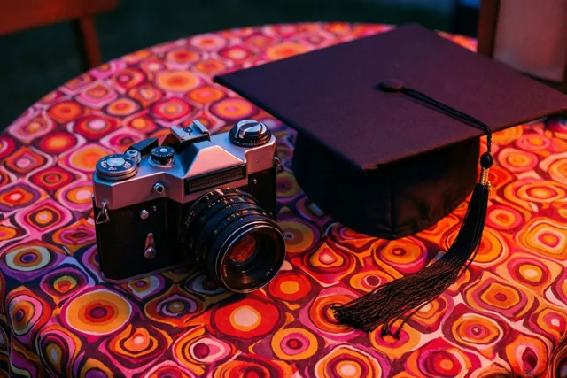 Een oude analoge camera en een afstudeerhoed liggen naast elkaar op een kleurrijk tafelkleed met concentrische cirkels in warm licht.