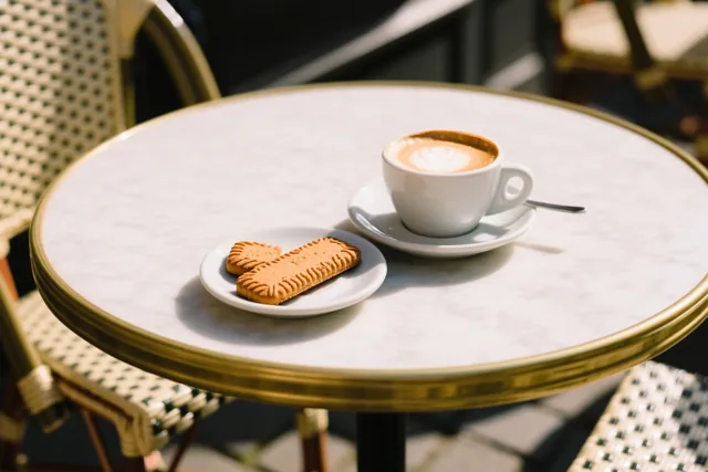 Een witte kop cappuccino met een figuurtje in het melkschuim en twee speculoosjes op een wit bordje staan op een ronde marmeren tafel met gouden rand op een zonnig terras met rieten stoelen.