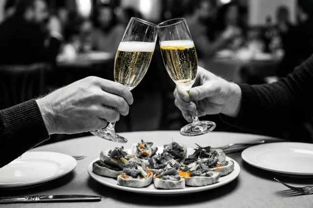 Twee handen klinken glazen bruisende cava boven een bord tapas, waarbij de cava en het eten in kleur zijn en de rest van het beeld zwart-wit is.