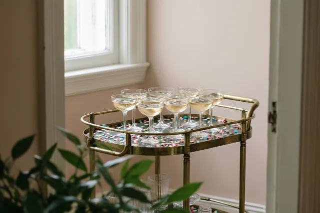 Op een oude messing serveerwagen staan champagnecoupes met bubbels tussen kleurrijke confetti, bij een raam met zacht daglicht.