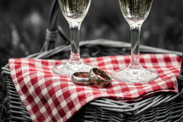Twee champagneglazen en twee versleten trouwringen liggen op een rood-wit geruite doek in een rieten picknickmand buiten, met een wazige natuurlijke achtergrond.