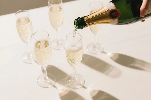 Een hand schenkt champagne uit een groene fles in een champagneglas, naast vier gevulde glazen op een witte tafel in warm zonlicht.