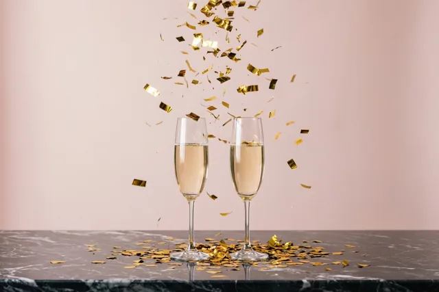 Twee champagneglazen met bruisende drank staan op een donkere marmeren tafel met vallende gouden confetti tegen een zachtroze achtergrond.