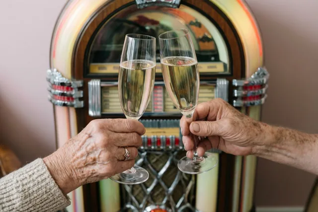 Twee oudere handen klinken met champagnefluiten voor een oude jukebox in warm licht, met een zachtroze muur op de achtergrond.