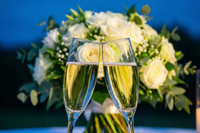 Twee champagneglazen met mousserende wijn staan schuin naar elkaar toe voor een onscherp wit bruidsboeket tegen een diepblauwe achtergrond.