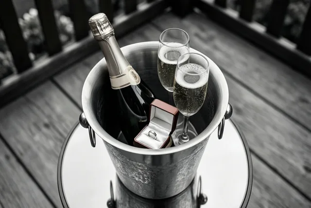 Een zilveren champagnekoeler met een fles champagne, twee gevulde glazen en een open ringdoosje met een ring staat op een tafel met glazen blad op een houten terras met een donkere reling.