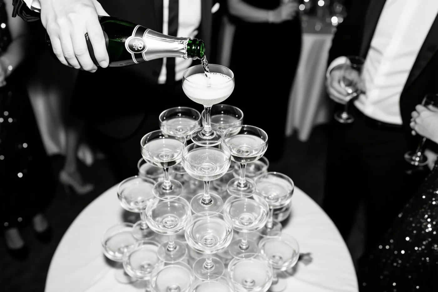 Een zwart-witfoto toont een hand die champagne schenkt in het bovenste glas van een piramide van champagneglazen op een formeel feest.