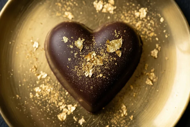 Een glanzend hart van donkere chocolade ligt op een gouden bord, met eetbaar bladgoud op de chocolade en het bord in warm licht.