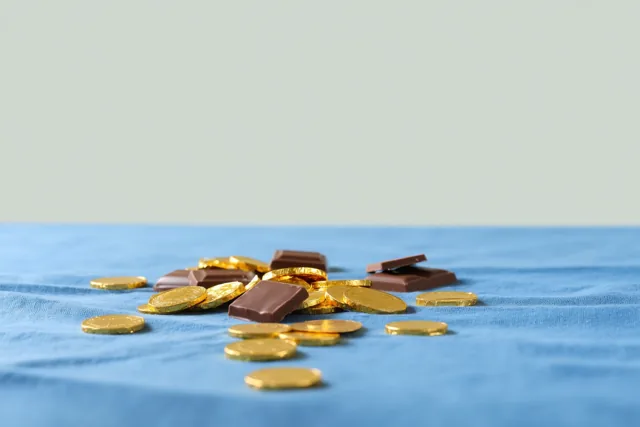 Chocolademunten in goudfolie en stukken gebroken chocoladereep liggen op een blauwe doek met zichtbare structuur tegen een effen lichtgroene achtergrond.