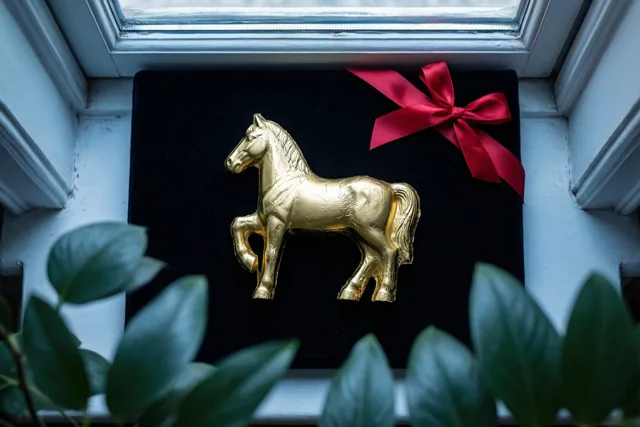 Een chocoladepaard in goudfolie met een rood lint staat op zwart fluweel op een vensterbank, met groene bladeren op de voorgrond en daglicht door het raam.