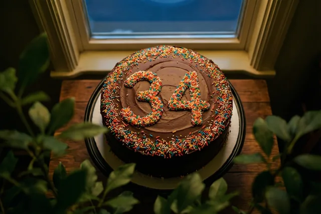 Een ronde chocoladetaart met 34 van gekleurde suikerstrooisels staat op een houten tafel bij een raam, omringd door groen.