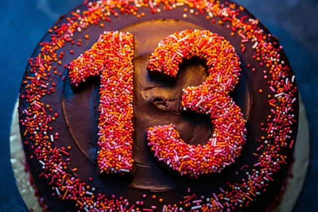 Een ronde chocoladetaart met 13 van gekleurde suikerstrooisels en een rand van rode, oranje en roze suikerstrooisels is van bovenaf te zien.