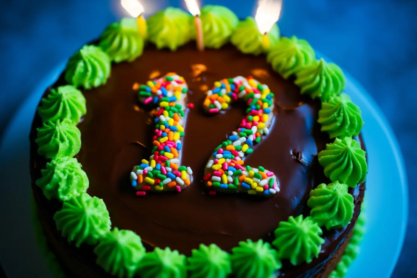 Van bovenaf is een ronde chocoladetaart te zien met het getal 12 van gekleurde suikerstrooisels en een groene gespoten glazuurrand, tegen een blauwe achtergrond met brandende kaarsjes bovenaan.