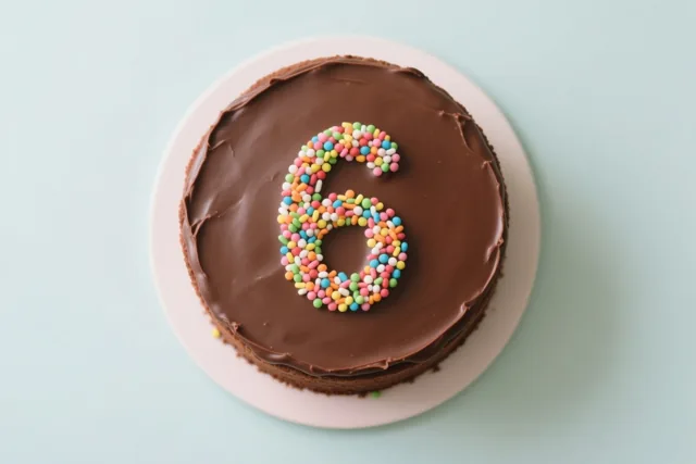 Een ronde chocoladetaart met glad glazuur en een 6 van gekleurde suikerstrooisels ligt op een wit bord tegen een lichtblauwe achtergrond, van bovenaf gezien.