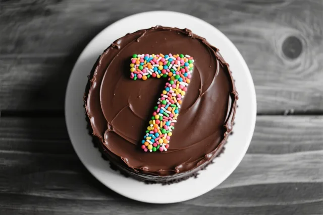 Een ronde chocoladetaart met glad glazuur en een 7 van gekleurde suikerstrooisels ligt op een wit bord tegen een donker houten achtergrond, van bovenaf gezien.