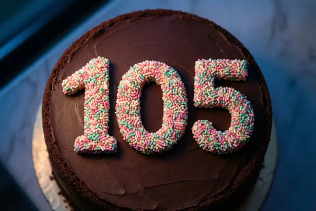Van bovenaf is een ronde chocoladetaart te zien met het getal 105 in gekleurde suikerstrooisels, op een witte kartonnen onderlegger tegen een blauwe achtergrond.