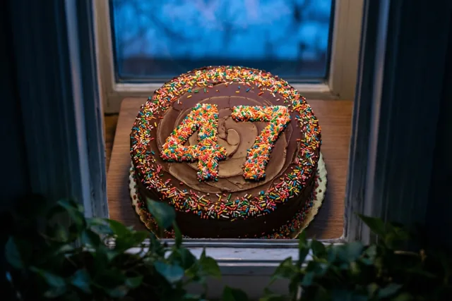 Een ronde chocoladetaart met 47 van gekleurde suikerstrooisels staat op een houten vensterbank, van bovenaf gezien tussen donkere gordijnen en groen met een blauwachtig uitzicht.