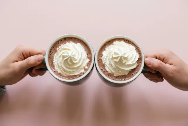 Twee handen klinken mokken warme chocolademelk met slagroom tegen elkaar voor een effen roze achtergrond.