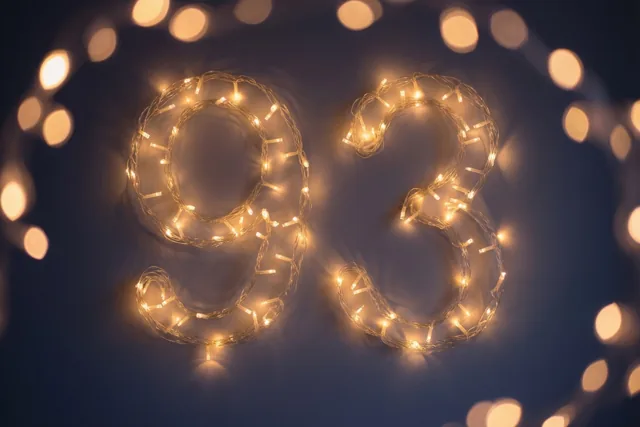 Warme lichtjes vormen het getal 93 tegen een donkere muur, met onscherpe lichtpunten op de achtergrond.