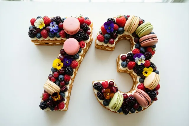 Een taart in de vorm van 73 staat op een effen witte ondergrond en is versierd met bessen, macarons en eetbare bloemen.