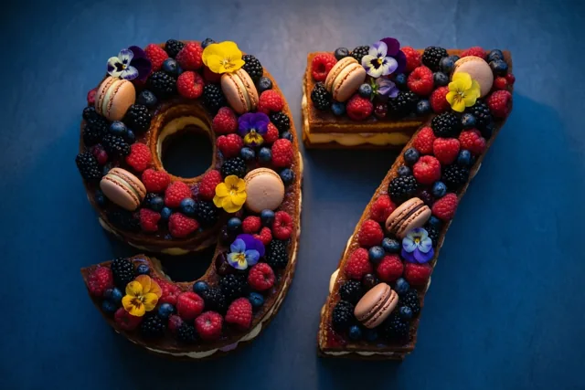 Een taart in de vorm van 97 is versierd met frambozen, bramen, blauwe bessen, macarons en eetbare bloemen tegen een effen donkerblauwe achtergrond.