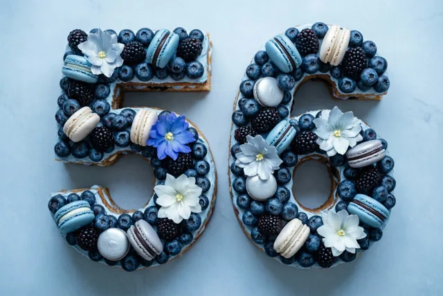 Een taart in de vorm van 56 is versierd met blauwe bessen, bramen, macarons en eetbare bloemen tegen een lichtblauwe achtergrond.