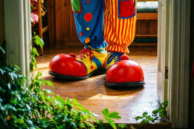 Door een deuropening zijn grote kleurrijke clownsschoenen met rode neuzen op een houten vloer te zien, met groen op de voorgrond.