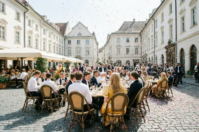 Op een druk caféterras aan een kasseiplein zitten gasten met hun rug naar de camera terwijl kleurrijke confetti valt, met historische gebouwen onder een heldere hemel op de achtergrond.