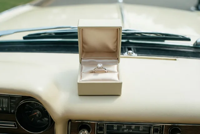 Een open crèmekleurig ringdoosje met een ring met één diamant staat op het dashboard van een crèmekleurige oldtimer in zacht licht.