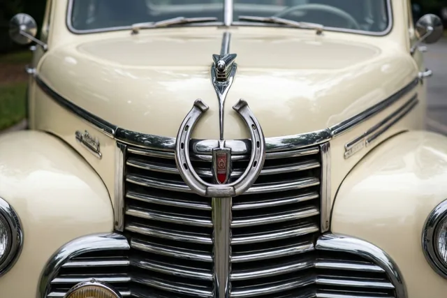 De grille van een crèmekleurige oldtimer is van dichtbij te zien met een groot verchroomd hoefijzer in het midden en glanzende accenten die de buitenomgeving weerspiegelen.