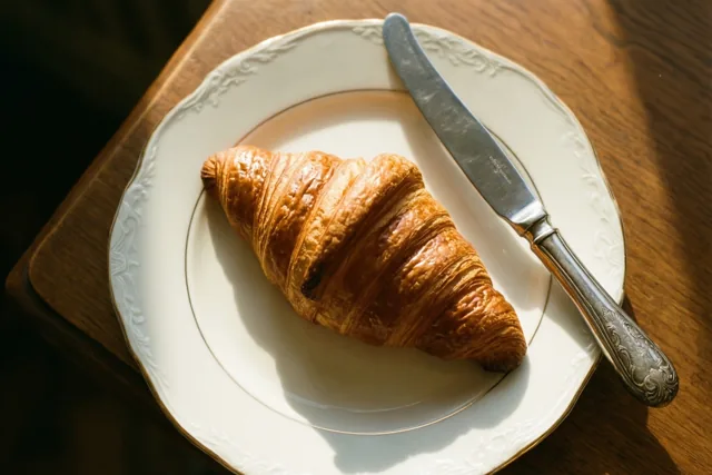 Een goudbruine croissant ligt op een wit porseleinen bord met een sierlijke rand, naast een zilveren botermes op een houten tafel in warm ochtendlicht.