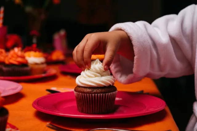 Een kinderhand uit een witte gewaadsmouw reikt naar een cupcake met wit glazuur op een roze bord, op een oranje tafelkleed met andere cupcakes op de achtergrond.