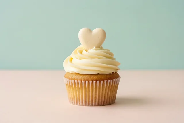 Een cupcake met witte crème en een hartje van glazuur staat op een zachtroze ondergrond tegen een licht muntgroene achtergrond.