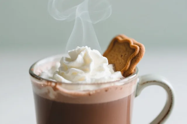 Een dampende mok chocolademelk met slagroom en een speculaasje is van dichtbij te zien tegen een zachte, neutrale achtergrond.