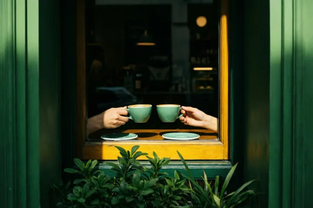 Twee handen houden bij een houten caféraam twee bijpassende groene koffiekopjes vast waaruit damp opstijgt, omringd door donkergroene raamkozijnen en bladeren, met zonlicht op de vensterbank.