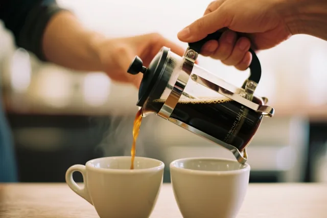 Een hand schenkt dampende koffie uit een cafetière in een van twee witte keramieken kopjes op een houten ondergrond, met warm licht en een wazige achtergrond.