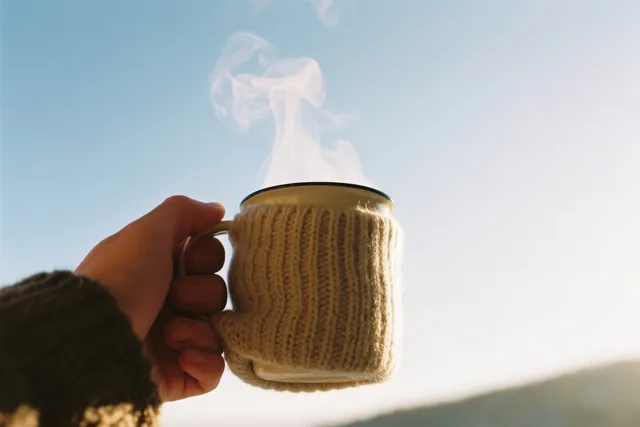 Een hand houdt een dampende mok met een beige gebreid hoesje vast tegen een heldere blauwe lucht.