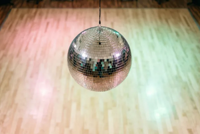 Een discobal hangt boven een lege houten dansvloer die door roze en groene lampen wordt verlicht.