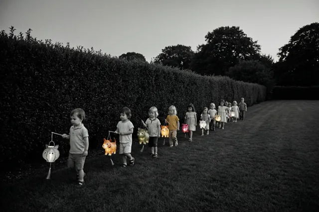 Kleine kinderen lopen in een rij langs een hoge haag met brandende dierenlantaarns die kleur brengen in de verder vrijwel monochrome schemering.