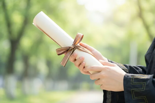 Een persoon in afstudeertoga houdt een opgerold diploma met een bruin lint vast tegen een onscherpe groene buitenachtergrond.