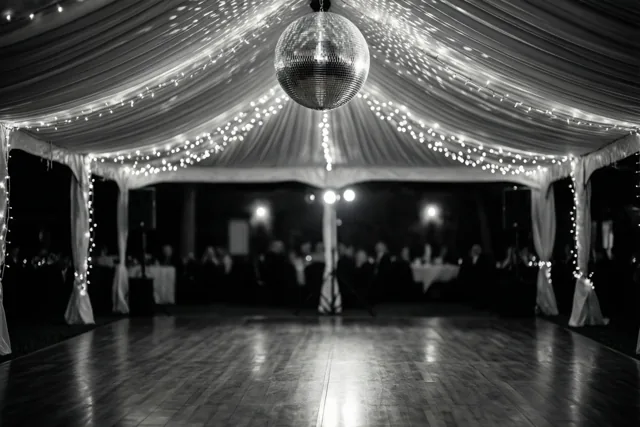 Een zwart-witfoto toont een lege houten dansvloer in een gedrapeerde feesttent met lichtslingers en een grote discobal die licht op de vloer weerkaatst.