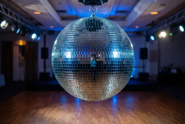 Een grote discobal hangt boven een glanzende houten dansvloer in een donkere feestzaal met blauw en warm licht.