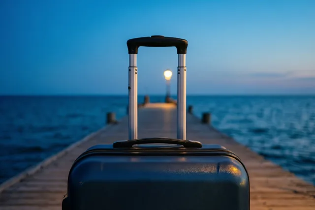 Een donkere koffer met uitgeschoven handvat staat bij schemering aan het einde van een houten pier, met in de verte een warm brandende straatlamp boven het blauwe water.