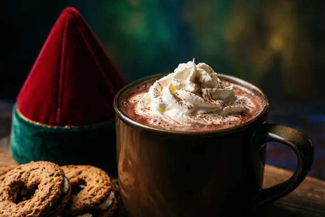 Een donkere mok warme chocolademelk met slagroom en cacaopoeder staat naast een rode fluwelen mijter en enkele pepernoten op een houten ondergrond, tegen een onscherpe groene en gouden achtergrond.