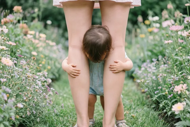 Een peuter met donker haar omhelst van achteren de benen van een vrouw in een weelderige tuin met kleurrijke bloemen.