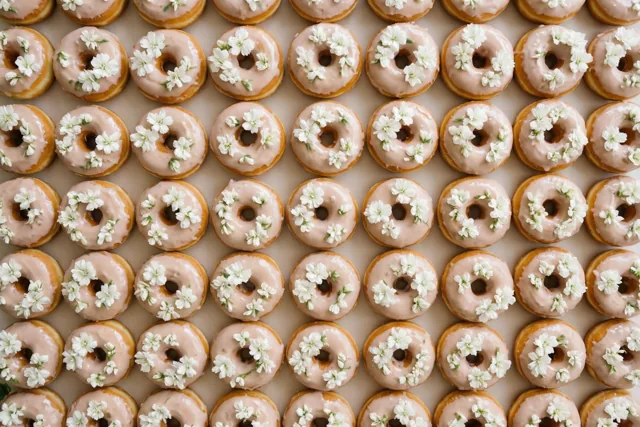 Geglazuurde donuts met kleine witte bloemen liggen van bovenaf gezien in nette rijen op een effen ondergrond in beige, lichtroze en witte tinten.
