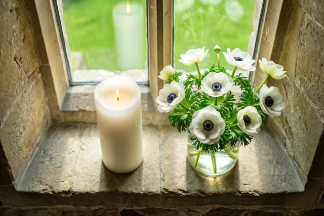 Een brandende witte doopkaars en een vaas witte anemonen staan op een zonnige stenen vensterbank in een kerk, met groen buiten het raam.