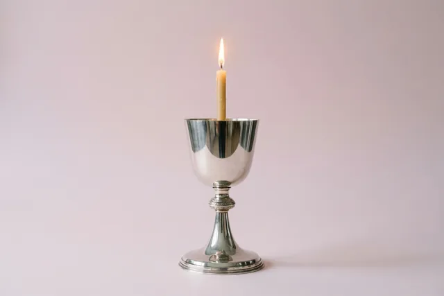 Een brandende doopkaars staat in een zilveren doopbekertje tegen een effen lichtroze achtergrond en werpt een warme gloed op het metaal.