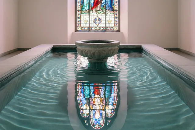 Een stenen doopvont staat in een rechthoekig waterbassin met rimpelend blauwgroen water dat een gekleurd glas-in-loodraam weerspiegelt, in een sobere ruimte met bleke muren.